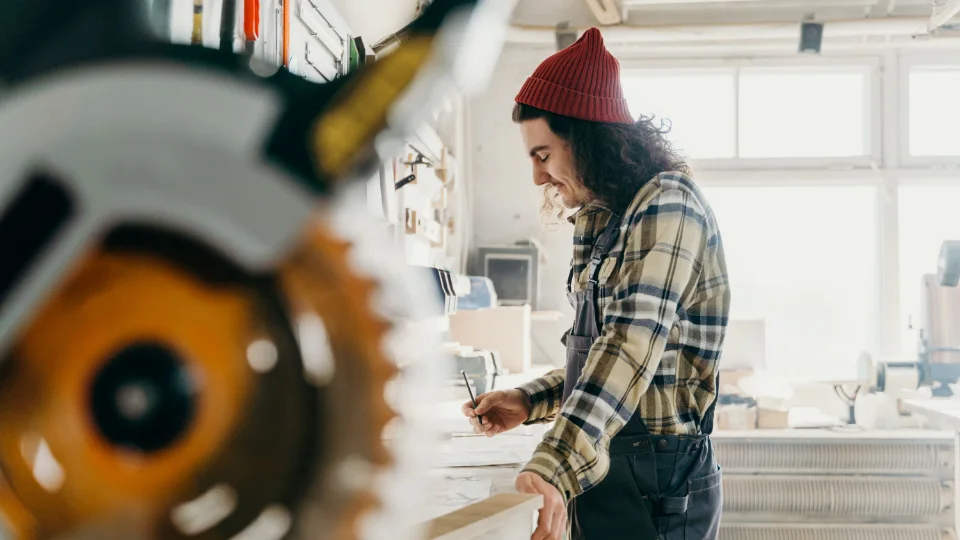 A carpenter measuring timber at a sunlit workshop bench